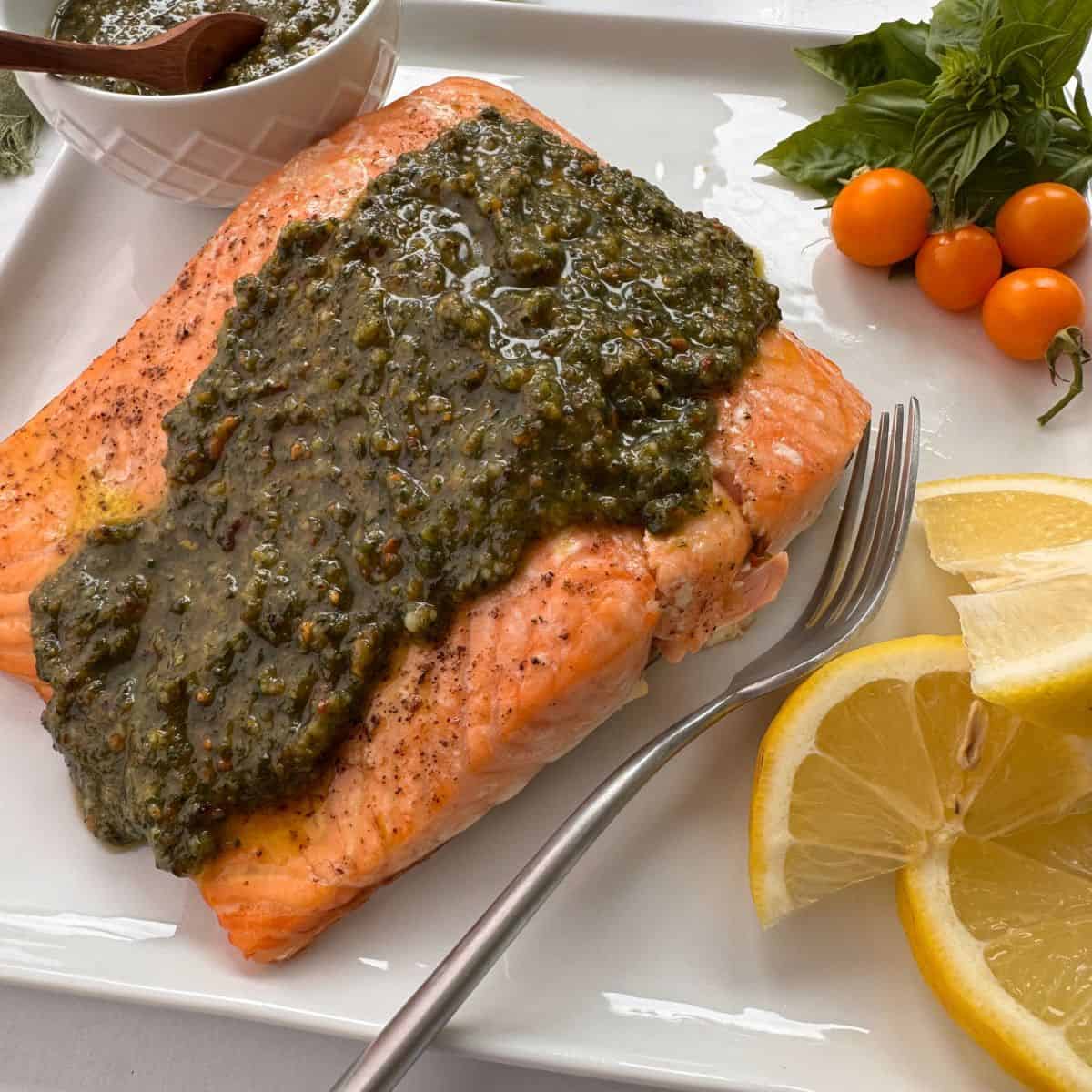 A salmon pesto fillet is served on a white plate with zesty lemon slices, a fork, fresh basil, cherry tomatoes, and a small bowl of extra sauce on the side.