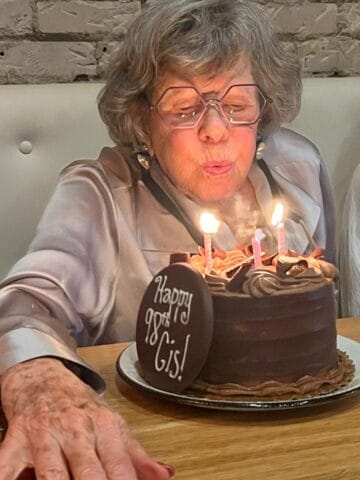 An elderly woman wearing glasses blows out candles on a chocolate birthday cake with a “Happy 98th Gigi!” plaque. The Sunday Thymes captured her joyful moment, seated at a table with a brick wall in the background.