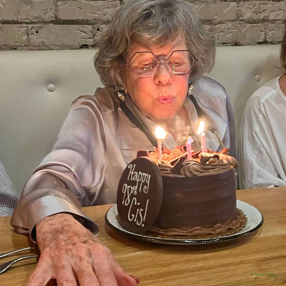 An elderly woman wearing glasses blows out candles on a chocolate birthday cake with a “Happy 98th Gigi!” plaque. The Sunday Thymes captured her joyful moment, seated at a table with a brick wall in the background.
