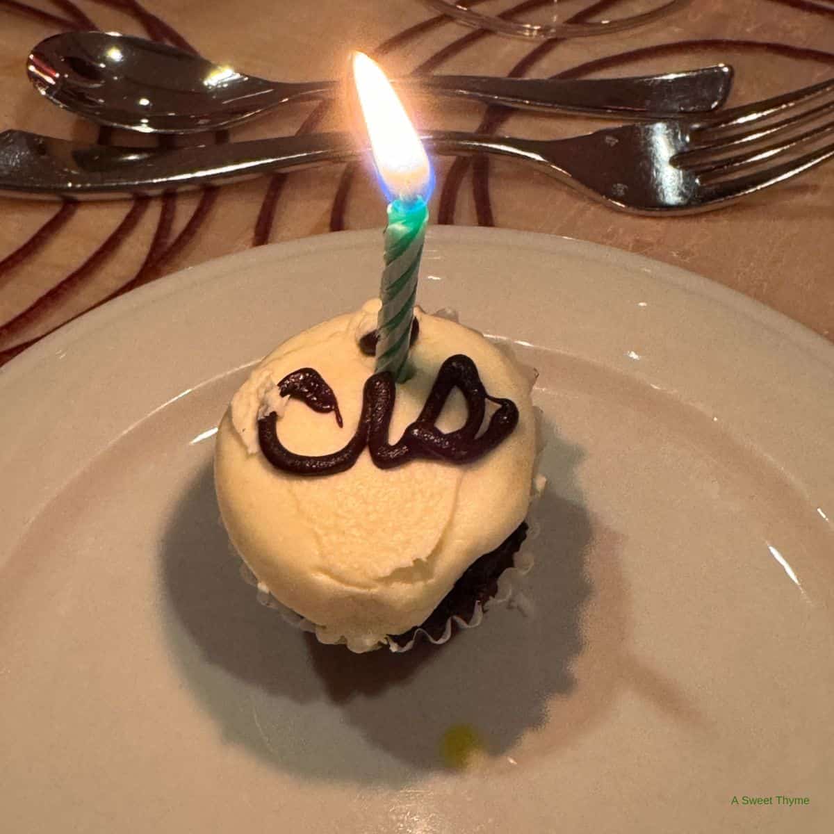 A cupcake with white frosting and a single lit candle sits on a plate, with chocolate writing on top. Spoons and a fork rest in the background on a Sunday Thymes tablecloth.