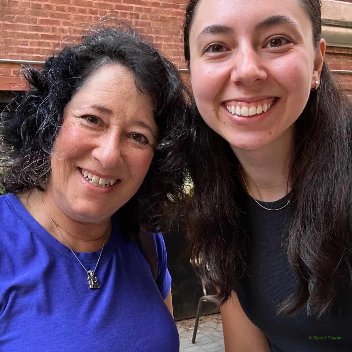 Two women smile outdoors, posing for a selfie on a sunny day. The woman on the left has curly dark hair and a blue shirt; the woman on the right has straight dark hair and a black top. A brick wall adds charm to their Sunday Thymes moment.