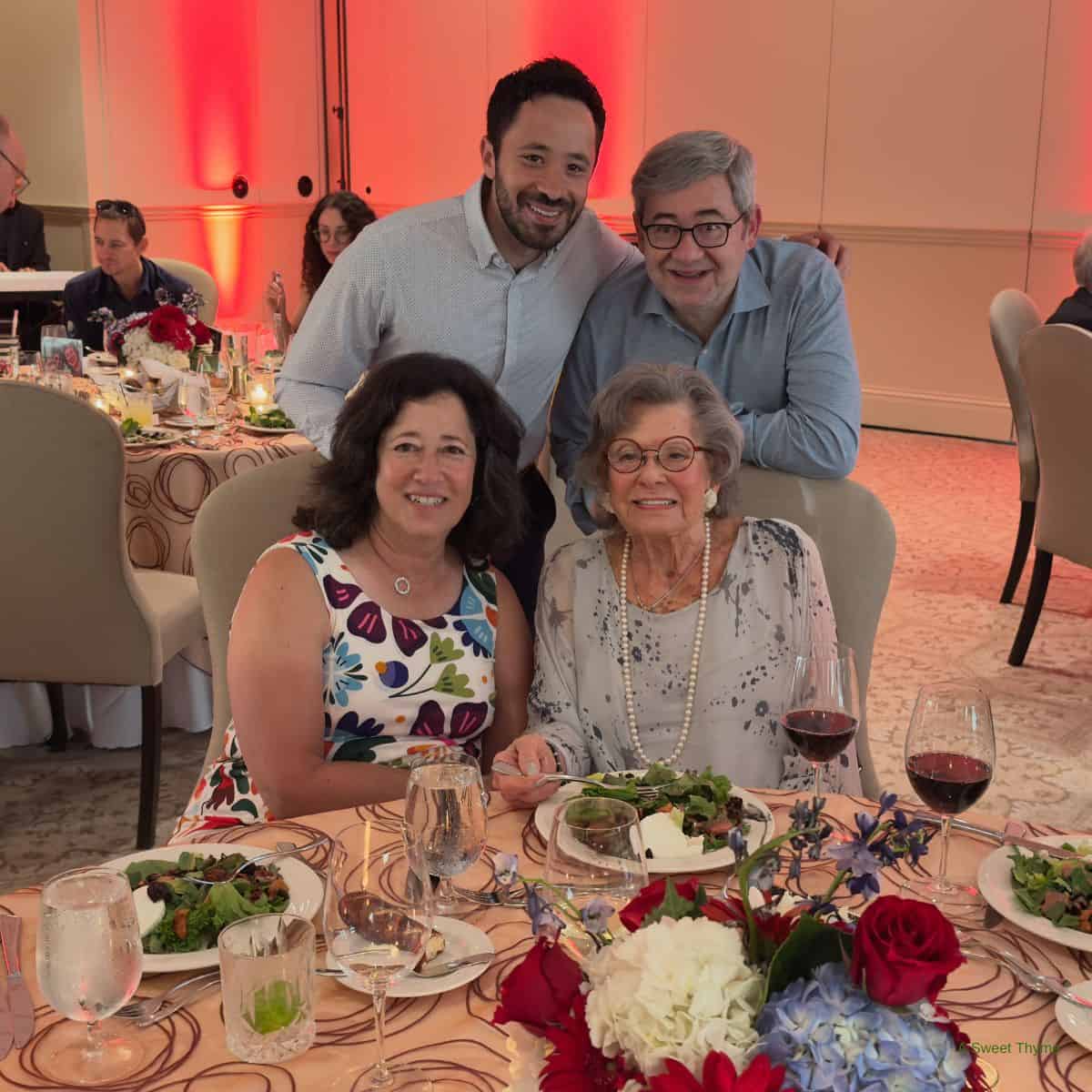 Four people smile at a banquet table with salads, wine, floral centerpieces, and a Sunday Thymes touch. Two women sit in front, two men stand behind. The background features guests, beige chairs, and orange-red uplighting.
