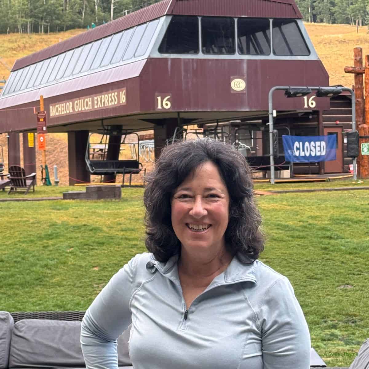 A woman with dark curly hair smiles while standing in front of the closed Bachelor Gulch Express 16 ski lift. The Sunday Thymes scene includes grassy slopes and trees in the background.