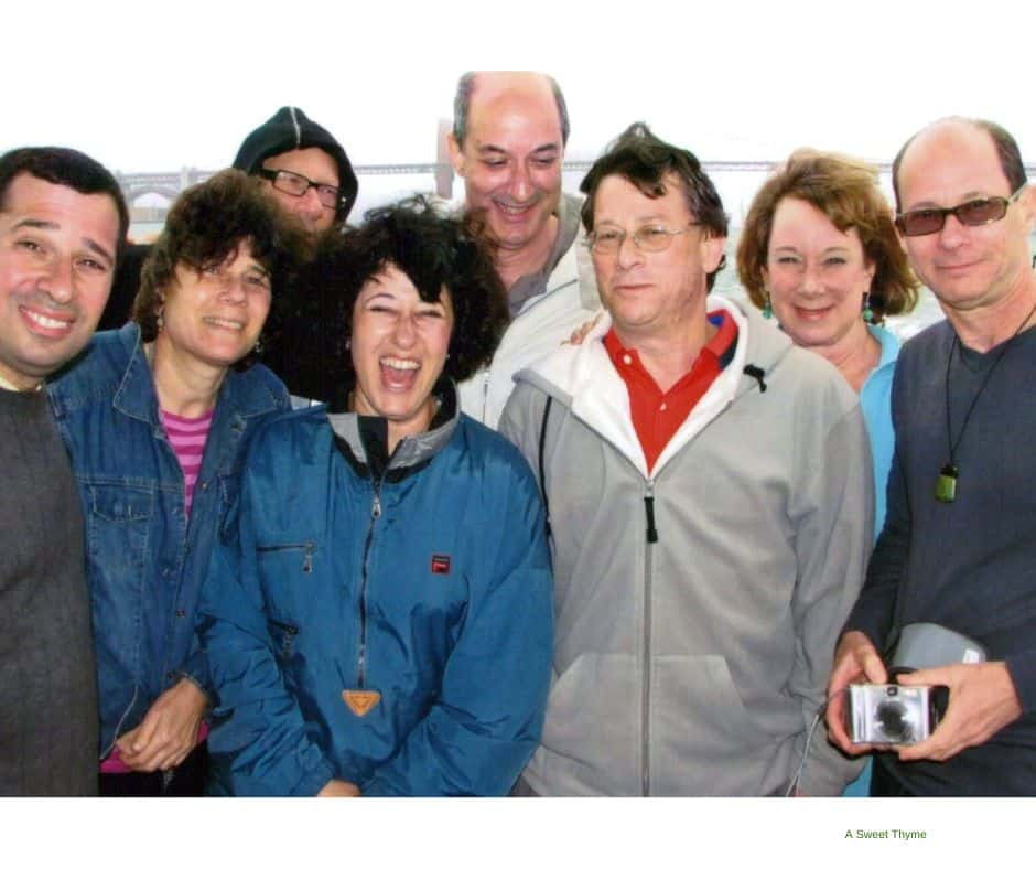 Nine adults, dressed in casual clothes, stand close together outdoors, smiling and laughing at the camera. One person holds a camera, capturing the joyful spirit of a Sunday Thymes outing with a bridge faintly visible in the background.