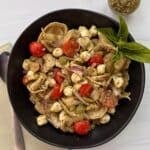 A black bowl filled with Tortellini Pesto Pasta Salad, cherry tomatoes, mozzarella balls, red onion slices, olives, and pesto, garnished with fresh basil. A spoon, fork, and napkin are beside the bowl, with extra pesto in a small dish nearby.