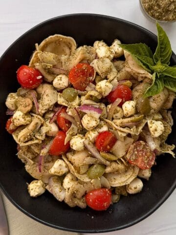 A black bowl filled with Tortellini Pesto Pasta Salad, cherry tomatoes, mozzarella balls, red onion slices, olives, and pesto, garnished with fresh basil. A spoon, fork, and napkin are beside the bowl, with extra pesto in a small dish nearby.