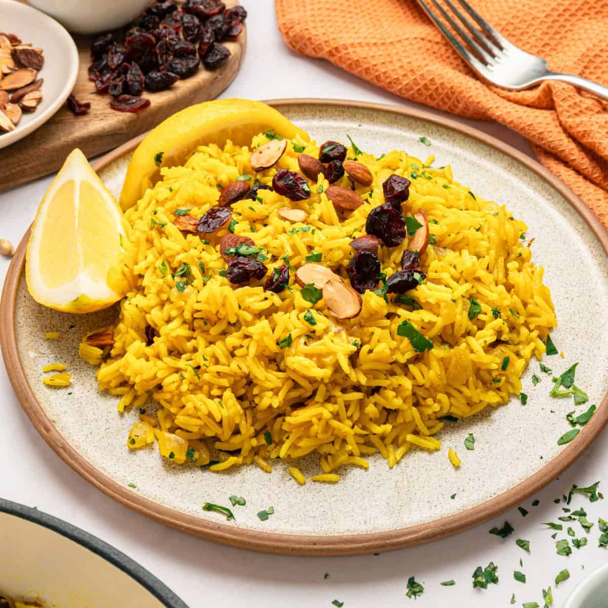 A plate of Mediterranean Rice, infused with turmeric and garnished with sliced almonds, dried cranberries, and chopped herbs, served with two lemon wedges. A fork, orange napkin, and a board with more almonds and cranberries are in the background.