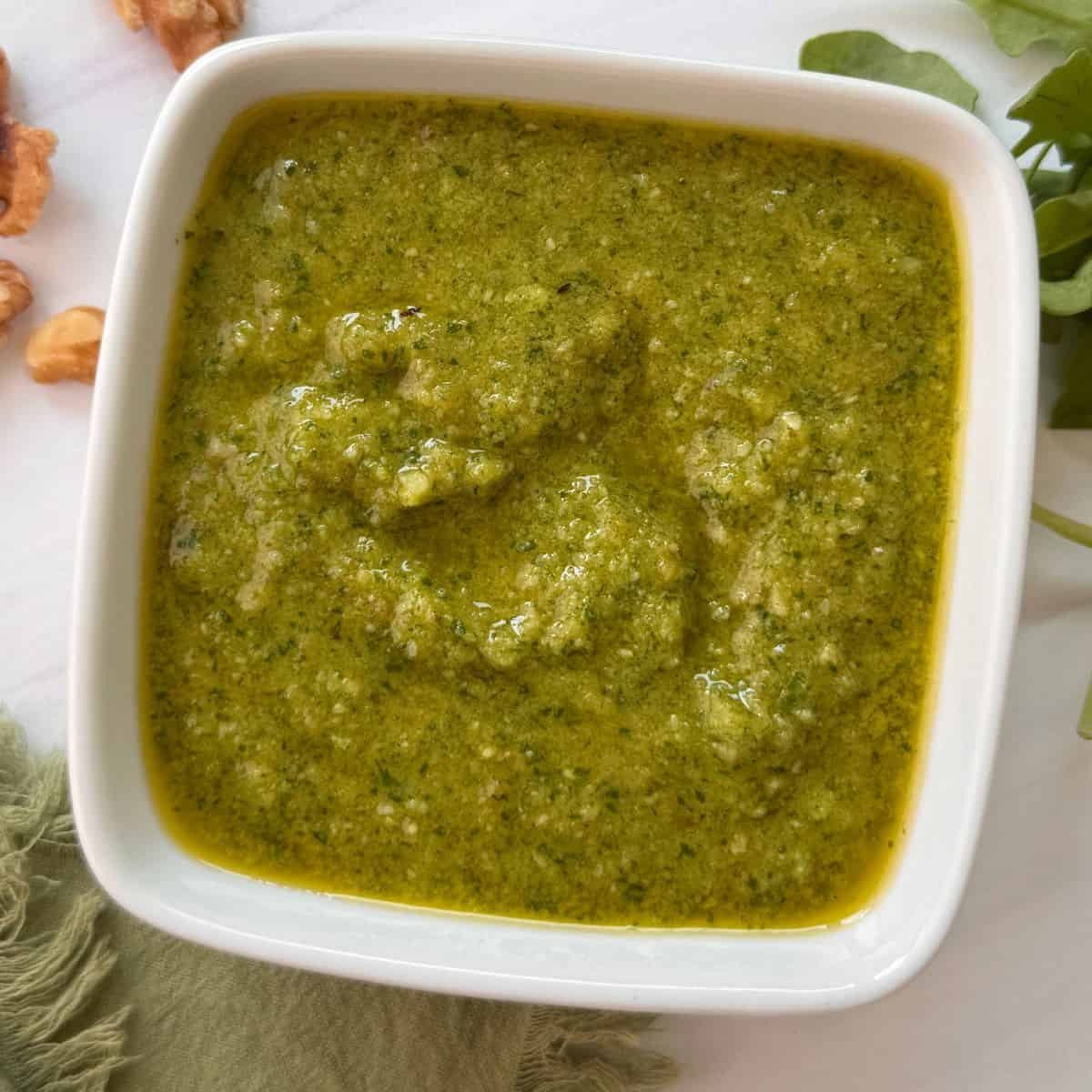 A square white bowl filled with walnut pesto sauce, its green, slightly chunky texture topped with a layer of olive oil. Fresh greens and walnut pieces are partially visible at the edges of the image.