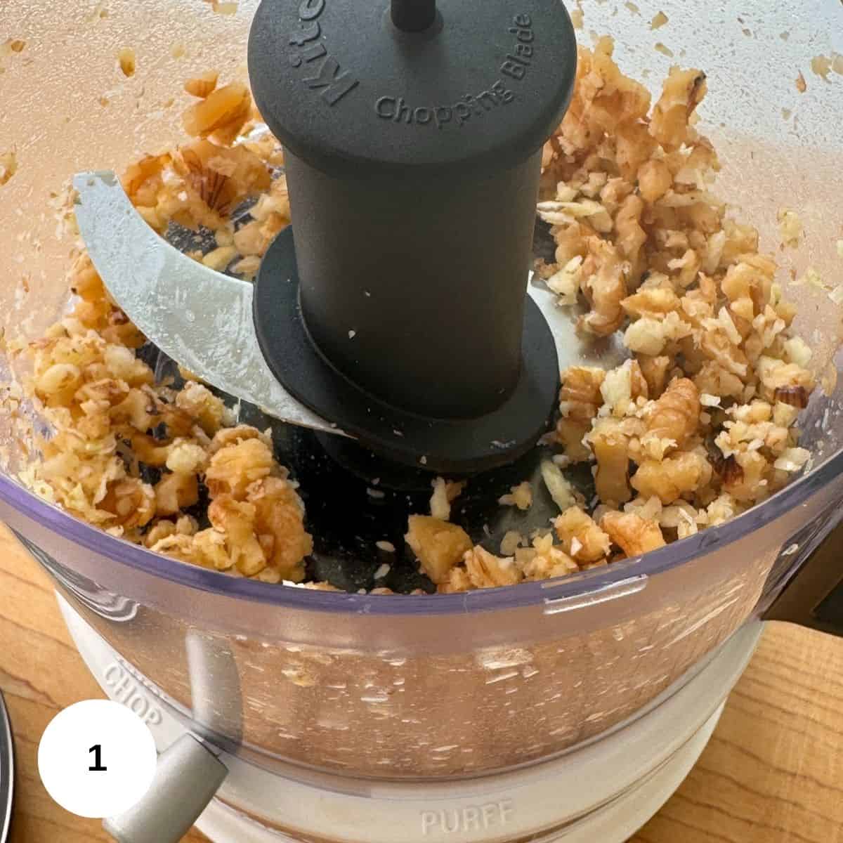Chopped nuts for walnut pesto are visible inside a food processor bowl with a sharp metal blade, sitting on a wooden countertop.