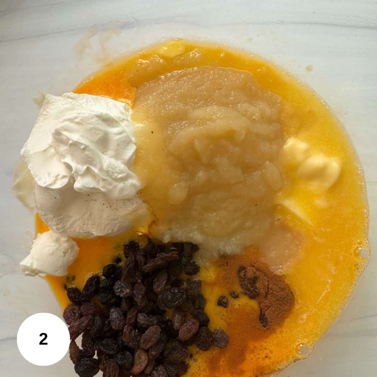 A bowl containing applesauce, whipped topping, melted butter, raisins, ground cinnamon, and vanilla-ingredients often found in Bundt Pan Noodle Kugel-rests on a white surface. A small white circle with the number 2 is in the bottom left corner.