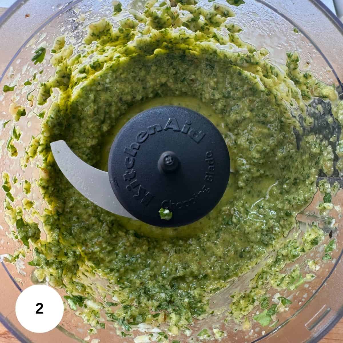 A close-up of a food processor containing freshly blended walnut pesto, with herbs visible around the sides and the blade in the center. The 
