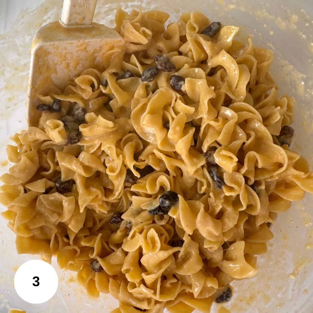 A close-up of creamy egg noodles and cinnamon raisins mixed in a bowl, evoking classic noodle kugel, with a white spatula partially visible on the left side.