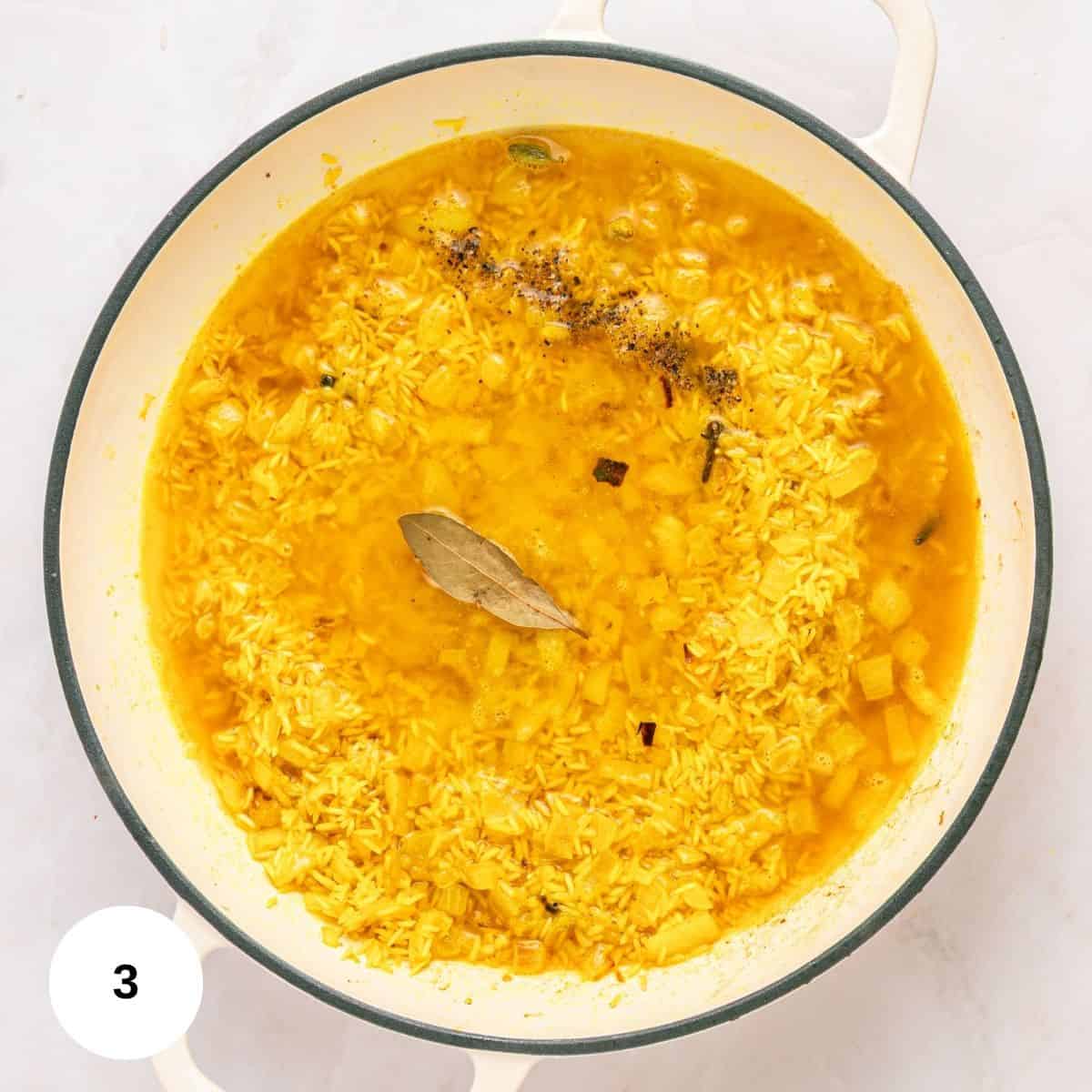 A pot filled with fragrant Mediterranean rice, golden from turmeric, simmers in broth with a bay leaf, ground black pepper, and visible spices. The pot rests on a light surface, with a white circle displaying the number 3 in the lower left corner.