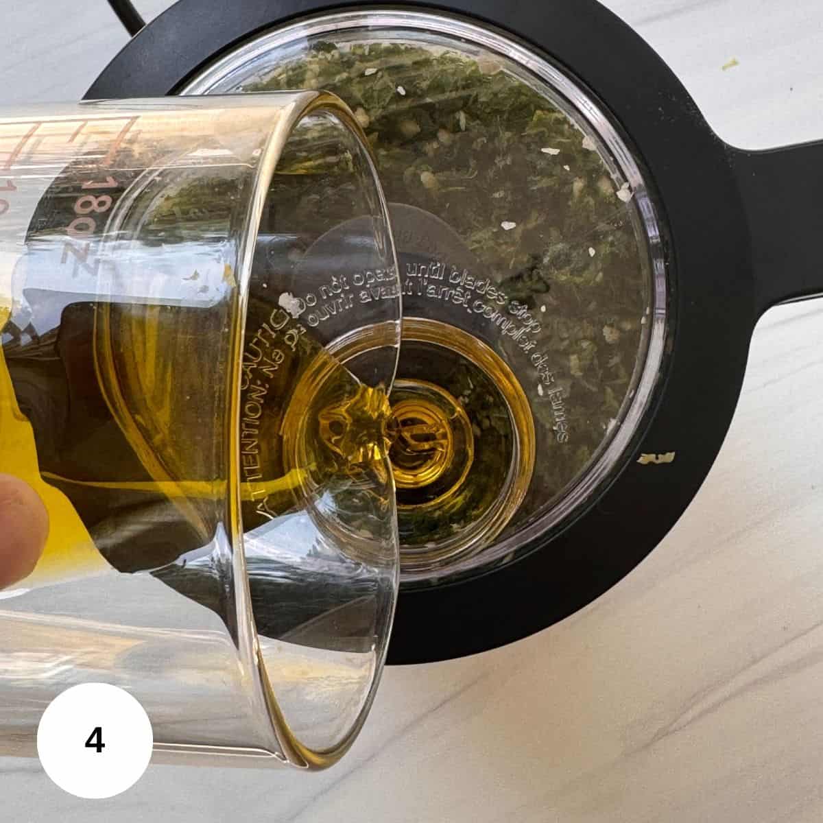 A hand pours oil from a measuring glass into a food processor containing chopped herbs for walnut pesto, viewed from above on a white surface.