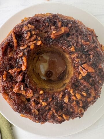 A round, baked monkey bread made in a Bundt Pan is topped with chopped nuts and cinnamon glaze, sitting on a white plate on a white surface next to a green napkin and a fork.