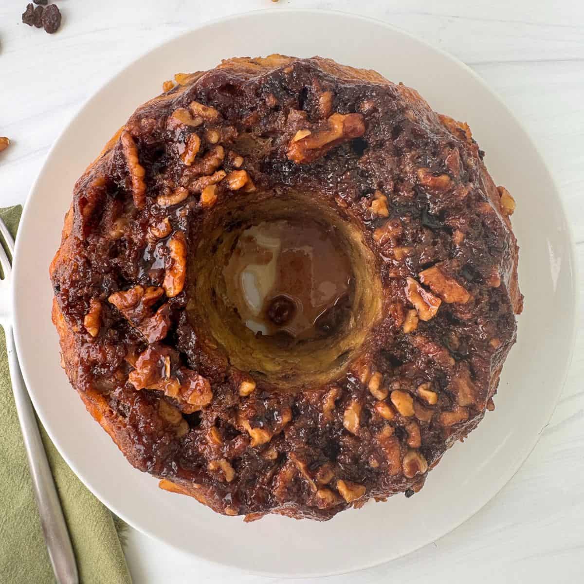 A round, baked monkey bread made in a Bundt Pan is topped with chopped nuts and cinnamon glaze, sitting on a white plate on a white surface next to a green napkin and a fork.