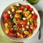 A bowl of gluten free gnocchi salad with cherry tomatoes, mozzarella balls, red bell pepper, and fresh basil sits on a light surface, accompanied by a spoon and green napkin.
