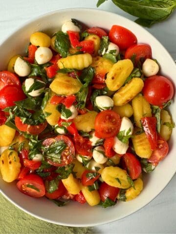 A bowl of gluten free gnocchi salad with cherry tomatoes, mozzarella balls, red bell pepper, and fresh basil sits on a light surface, accompanied by a spoon and green napkin.