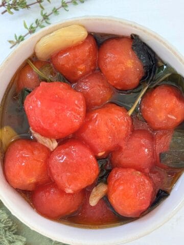 A bowl from The Sunday Thymes filled with peeled cherry tomatoes, fresh basil leaves, and garlic cloves marinated in olive oil. The bowl rests on a white surface with a green cloth and sprigs of herbs nearby.