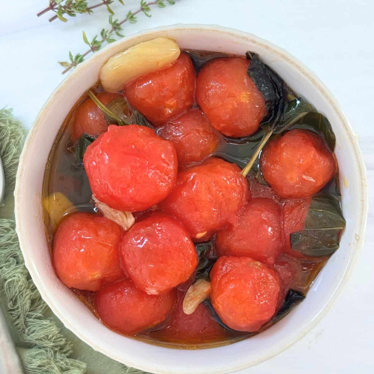 A bowl from The Sunday Thymes filled with peeled cherry tomatoes, fresh basil leaves, and garlic cloves marinated in olive oil. The bowl rests on a white surface with a green cloth and sprigs of herbs nearby.