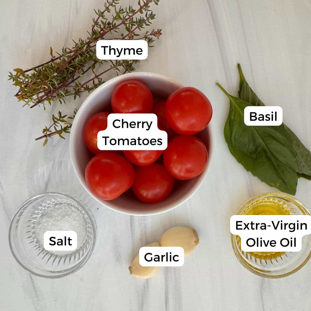 A bowl of cherry tomatoes is surrounded by thyme, basil leaves, a small glass dish of salt, two garlic cloves, and extra-virgin olive oil—classic Mediterranean ingredients for making cherry tomato confit—neatly labeled on a white surface.