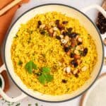 A pan of vibrant Mediterranean rice, colored with turmeric and garnished with chopped almonds, dried cranberries, and fresh parsley, sits beside an orange towel and a wooden spoon.