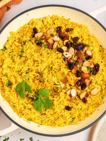 A pan of vibrant Mediterranean rice, colored with turmeric and garnished with chopped almonds, dried cranberries, and fresh parsley, sits beside an orange towel and a wooden spoon.