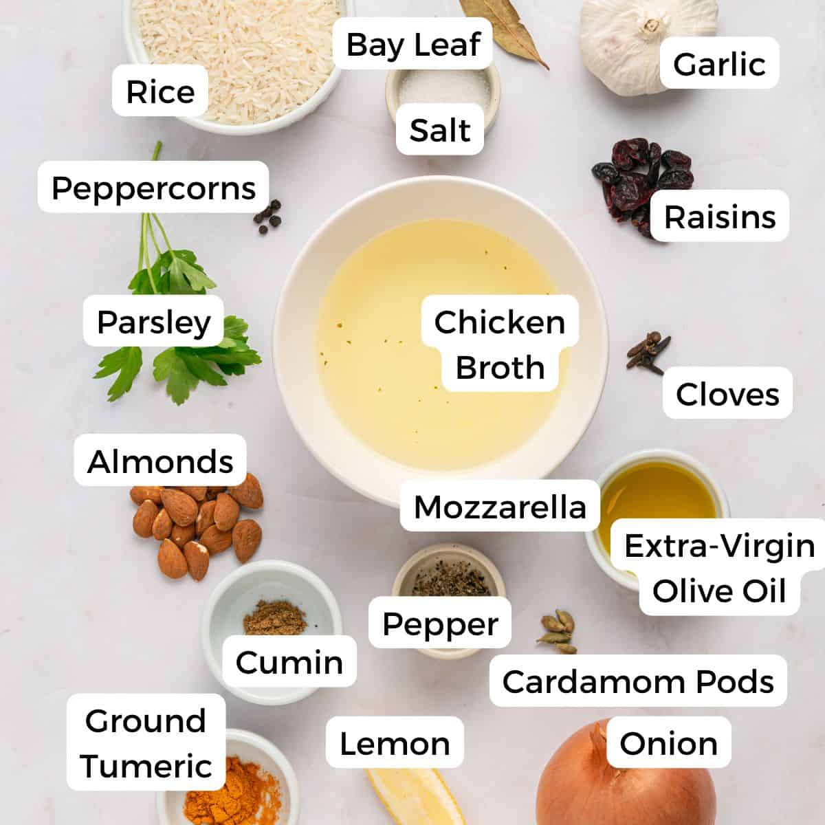 Top-down view of labeled ingredients for Mediterranean Rice, featuring rice, turmeric, almonds, bay leaf, garlic, peppercorns, parsley, chicken broth, raisins, spices like cumin and cardamom pods, lemon, onion, mozzarella, olive oil, salt and cloves.