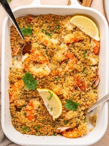 A white baking dish filled with shrimp oreganata, baked to perfection and topped with golden, crispy breadcrumbs, garnished with lemon wedges and parsley. Two serving spoons are in the dish, and a lemon sits beside it on the table.