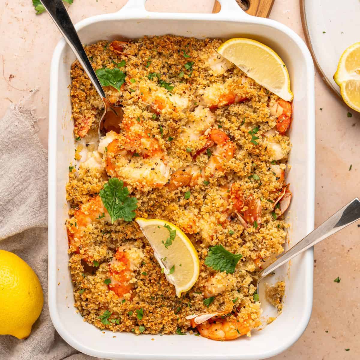 A white baking dish filled with shrimp oreganata, baked to perfection and topped with golden, crispy breadcrumbs, garnished with lemon wedges and parsley. Two serving spoons are in the dish, and a lemon sits beside it on the table.