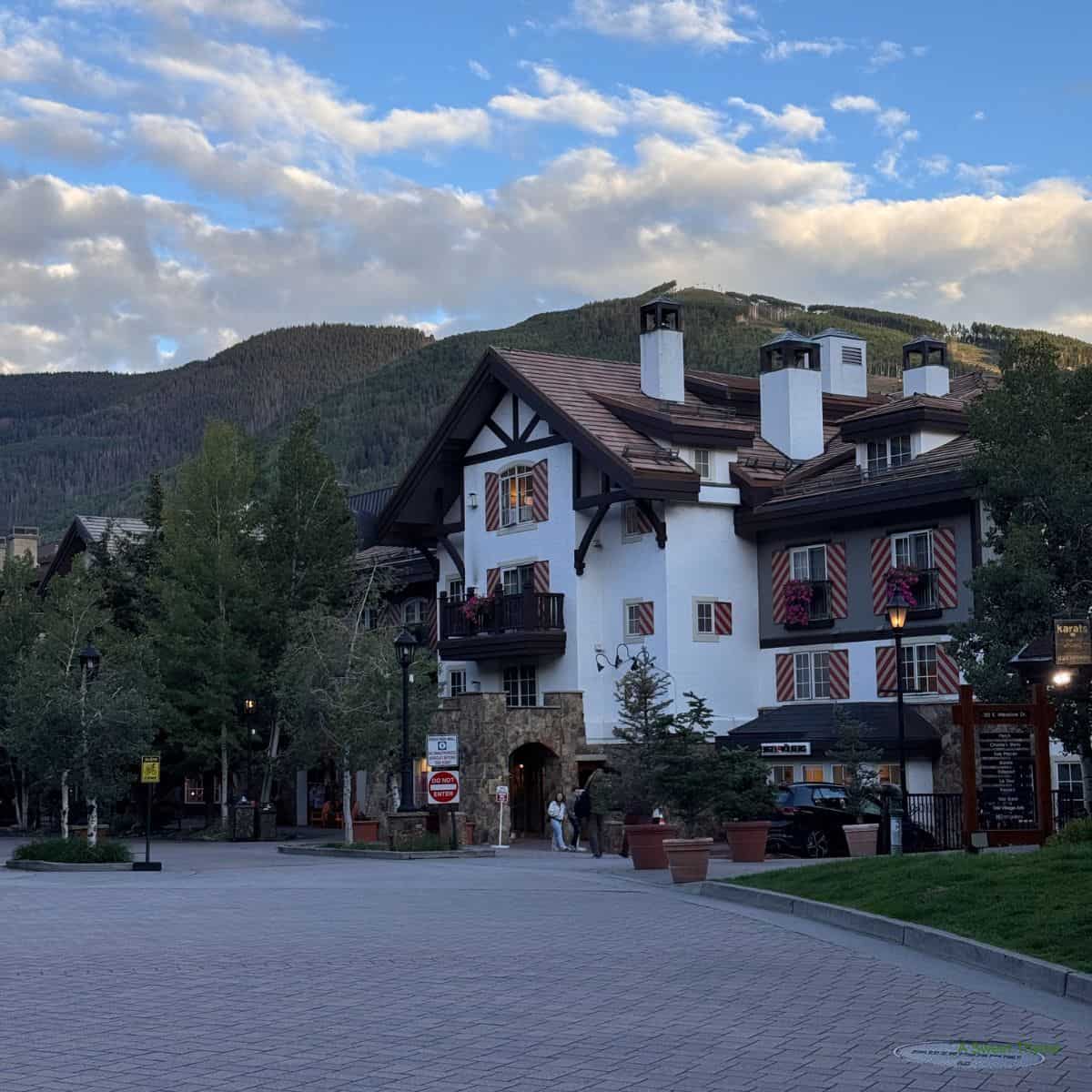 A picturesque alpine-style building with white walls and dark wooden beams stands near green trees and mountains under a partly cloudy sky. A few people stroll along the paved plaza in front, giving the scene a relaxed Sunday Thymes charm.