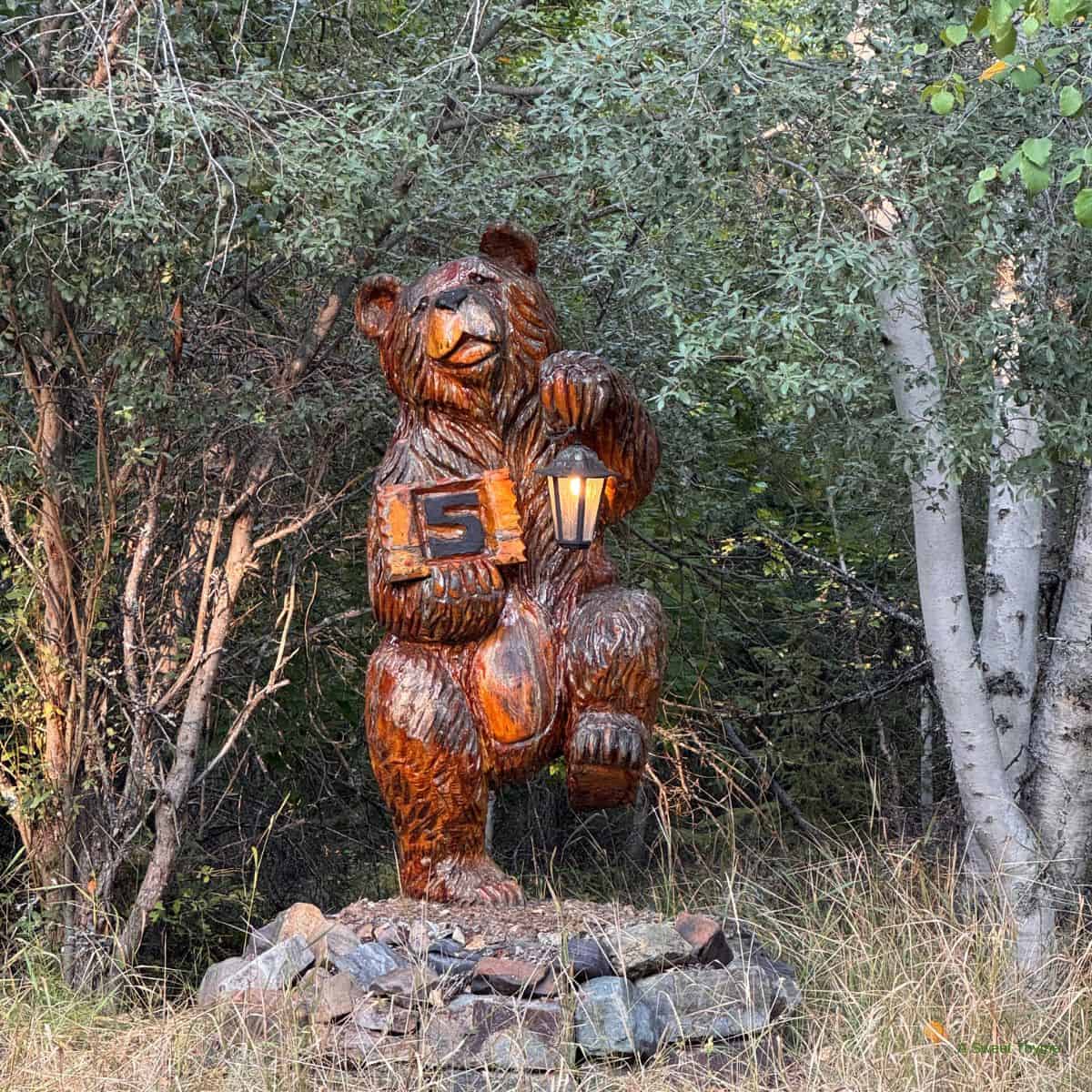A carved wooden bear statue stands on rocks in a forest, holding a lantern in one paw and a block with the number 5 in the other. The scene, perfect for The Sunday Thymes, features dense trees and tall grass in the background.