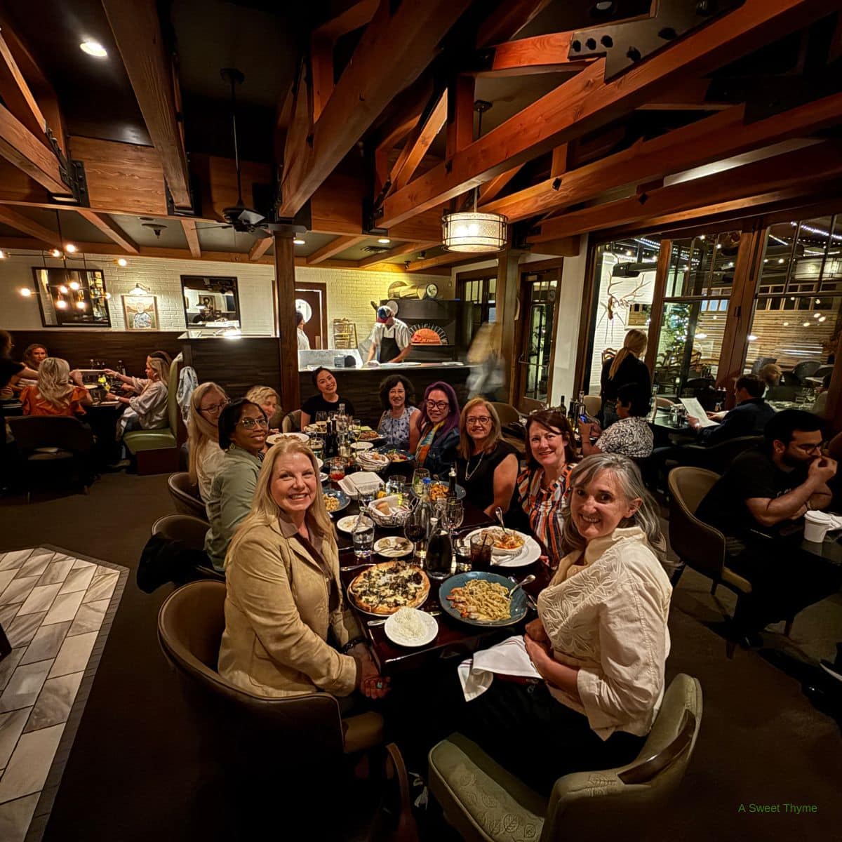 A group of women sit around a table at The Sunday Thymes restaurant, smiling at the camera. The table is filled with food and drinks, set against warm lighting, wooden beams, and other diners in the background.