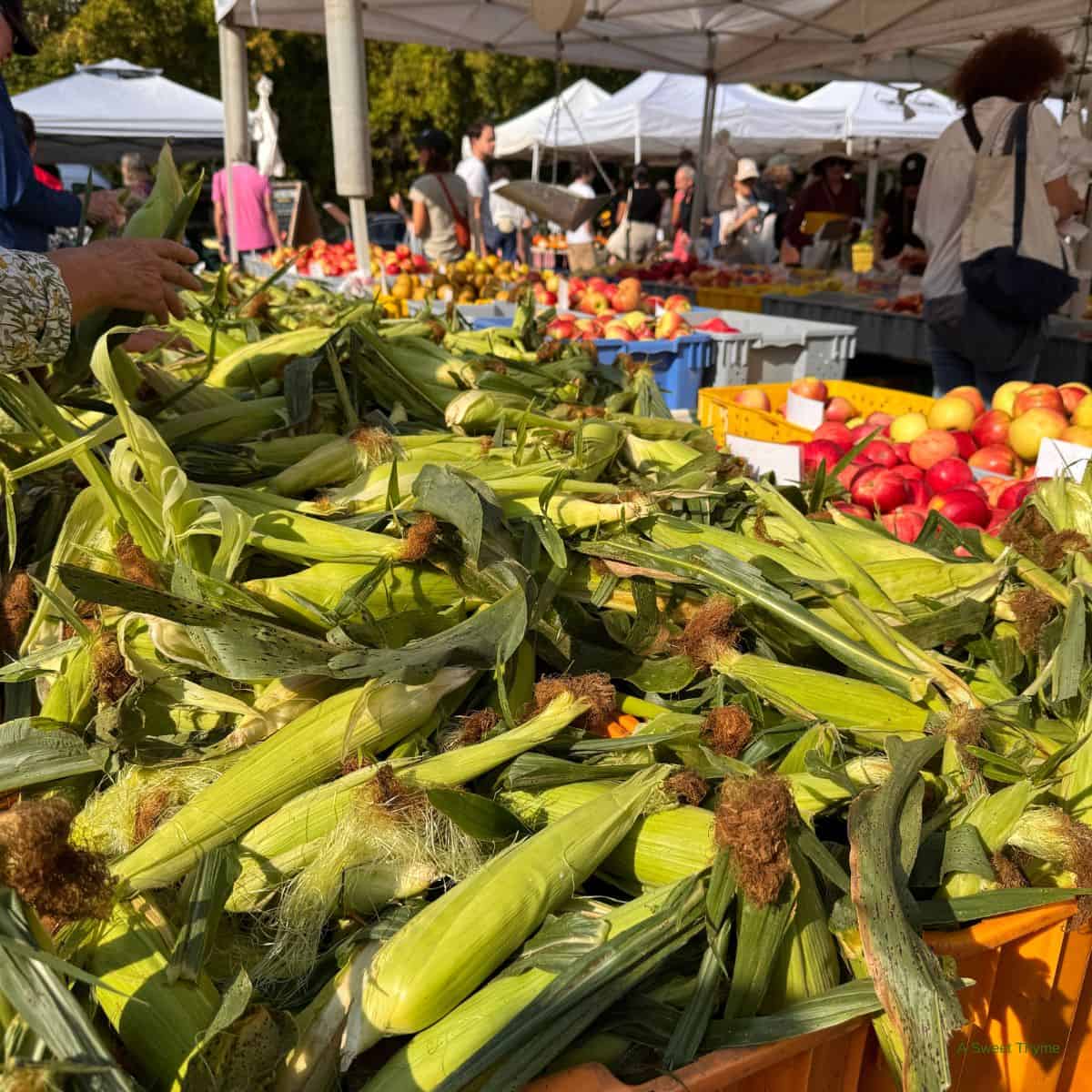 Fresh ears of corn with husks are piled high on a market table. In the background, people shop for fruits and vegetables under white canopy tents at an outdoor farmers market featured by The Sunday Thymes.