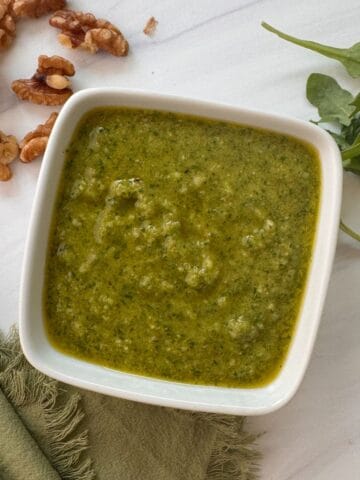 A square white bowl filled with green walnut pesto sauce sits on a white surface, with arugula leaves and walnut pieces nearby. A green cloth and silver spoon are also partially visible next to the bowl.