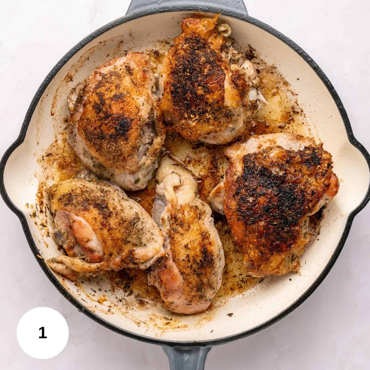 Five seasoned, browned chicken thighs cook in a round, white enameled skillet with a gray handle, sitting on a light surface. This one skillet meal makes the chicken appear crispy and perfectly cooked.
