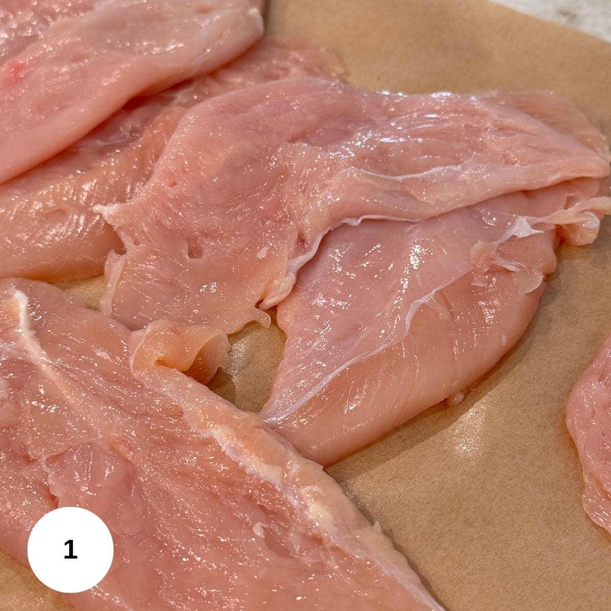 Raw chicken breast fillets laid out on brown parchment paper, ready for preparation-perfect for making classic Chicken Milanese. A white circle with the number 1 appears in the bottom left corner of the image.