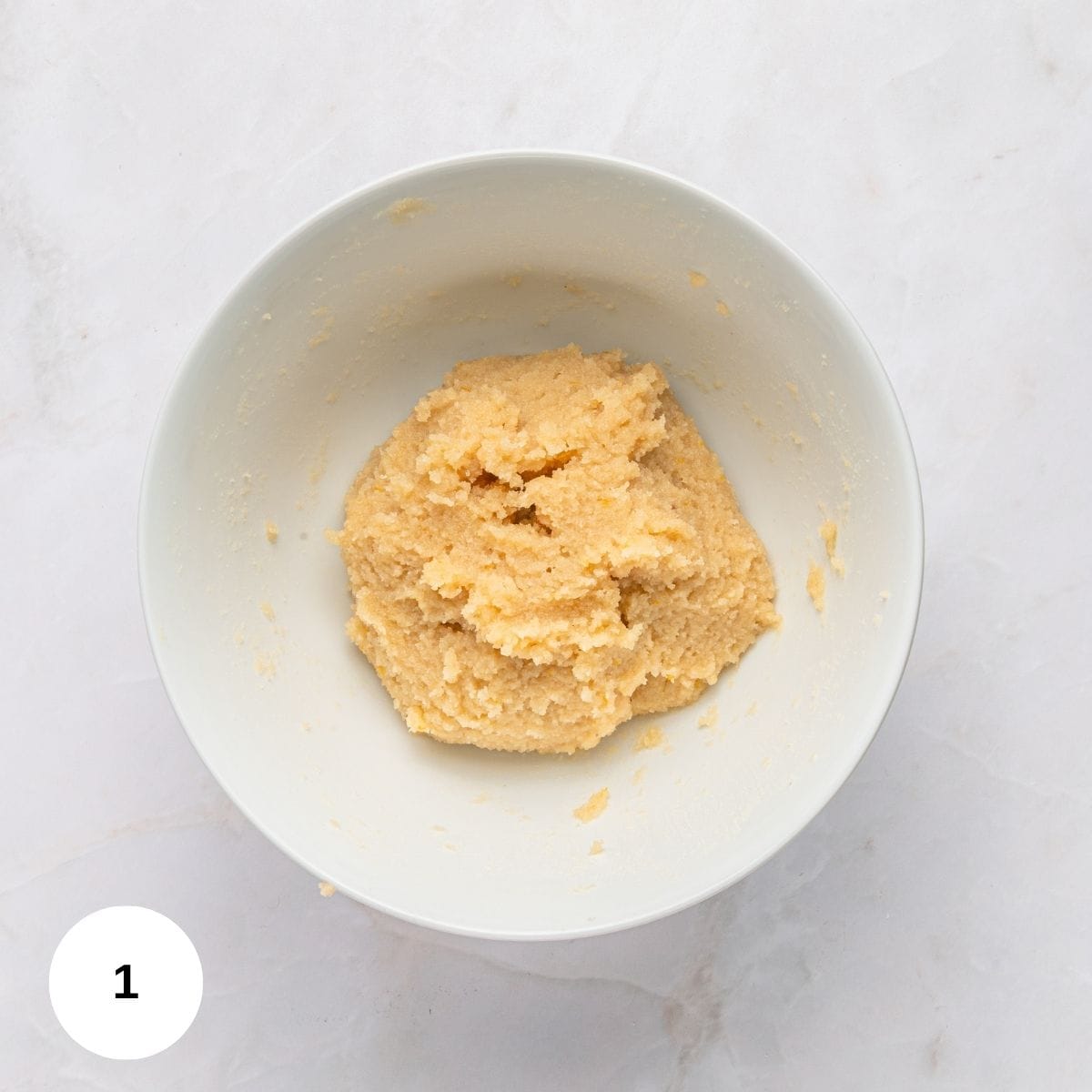 A white bowl filled with a thick, creamy, pale yellow mixture for Italian almond cookies with almond flour sits on a light marble surface. A white circle with the number 1 is in the bottom left corner.