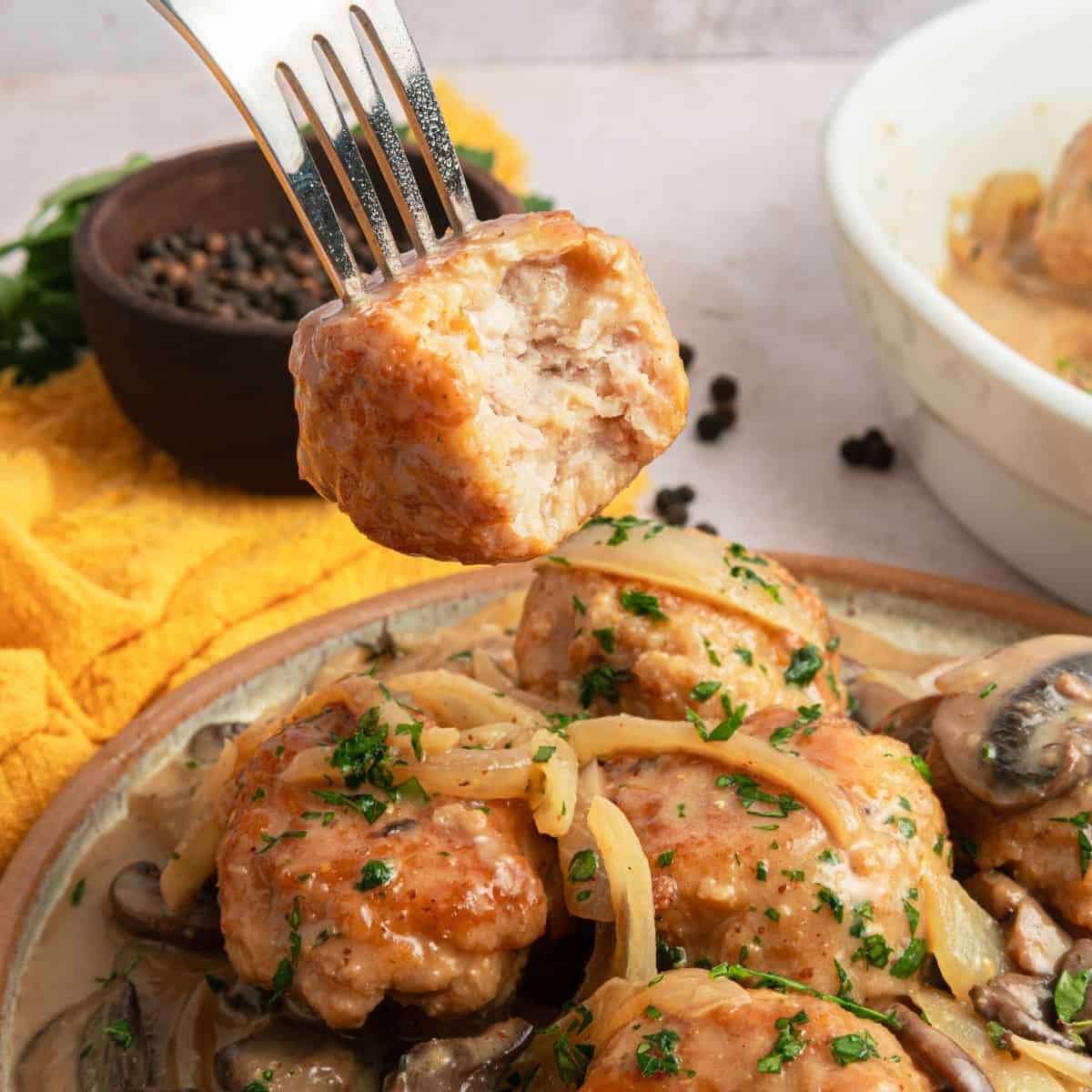 A fork holds a bite of Chicken Marsala Meatballs topped with sauce, above a plate of gluten free meatballs garnished with herbs and onions, with mushrooms on the side. A yellow cloth and a bowl of peppercorns are in the background.