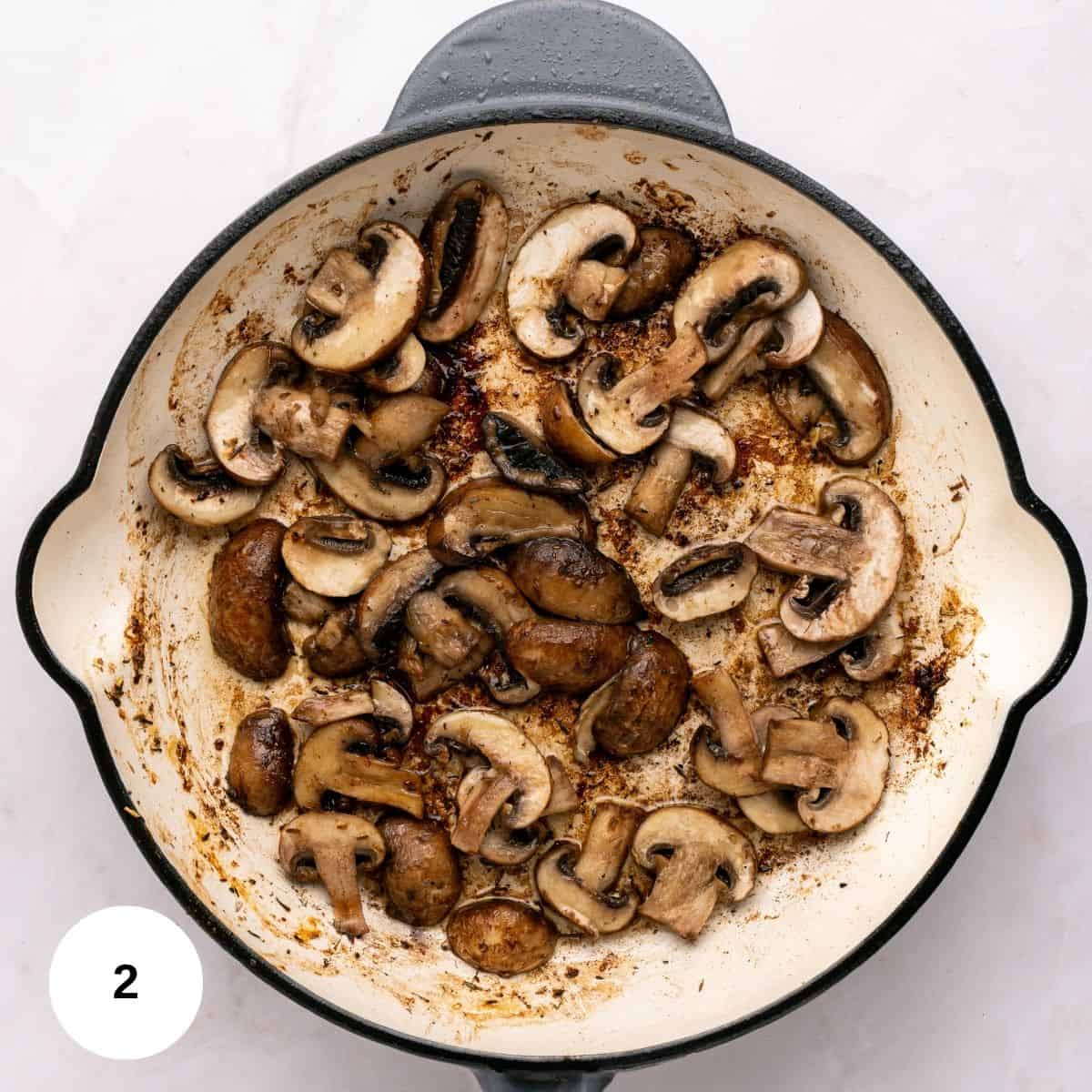 Sliced mushrooms being sautéed in a white skillet, showing browning and caramelization-perfect for Chicken Marsala. The skillet rests on a light-colored surface, with a small white circle marked 