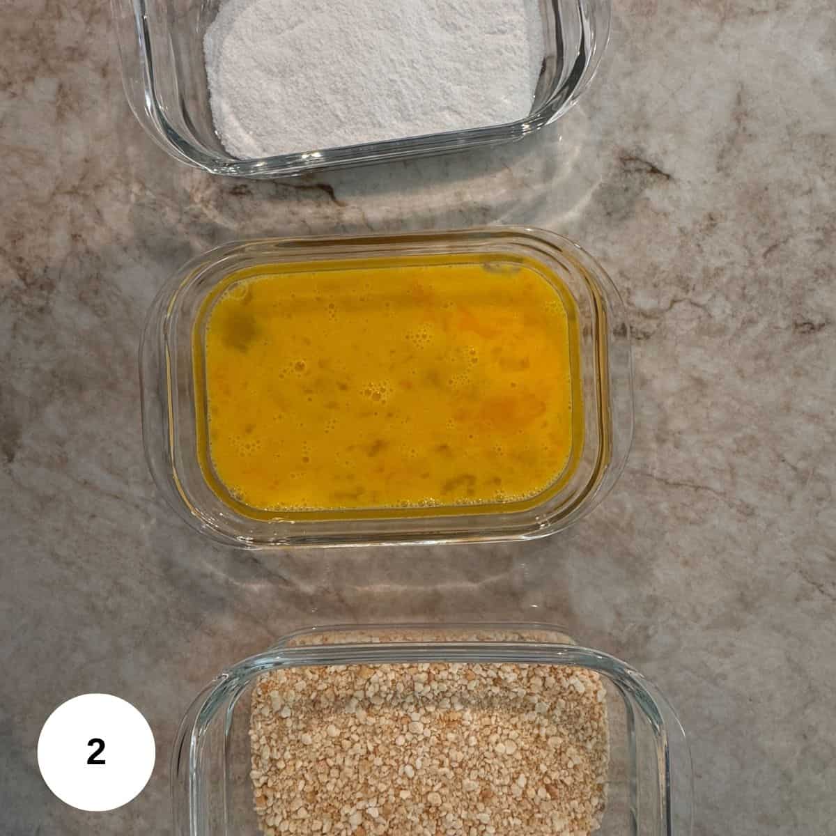 Three glass containers on a marble surface: the top holds gluten-free flour, the middle has beaten eggs, and the bottom contains breadcrumbs. A white circle with the number 2 is in the bottom left corner-perfect for Chicken Milanese prep.