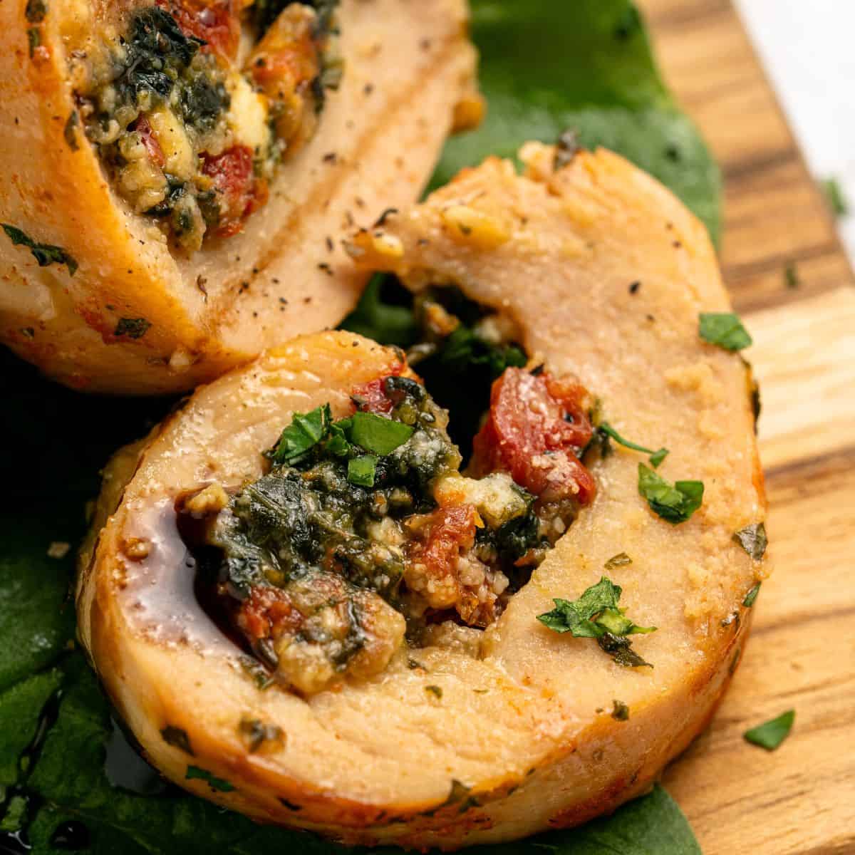 Close-up of two slices of chicken roulade filled with spinach, sundried tomatoes, and cheese, garnished with chopped herbs and served on a wooden board with fresh green leaves underneath.