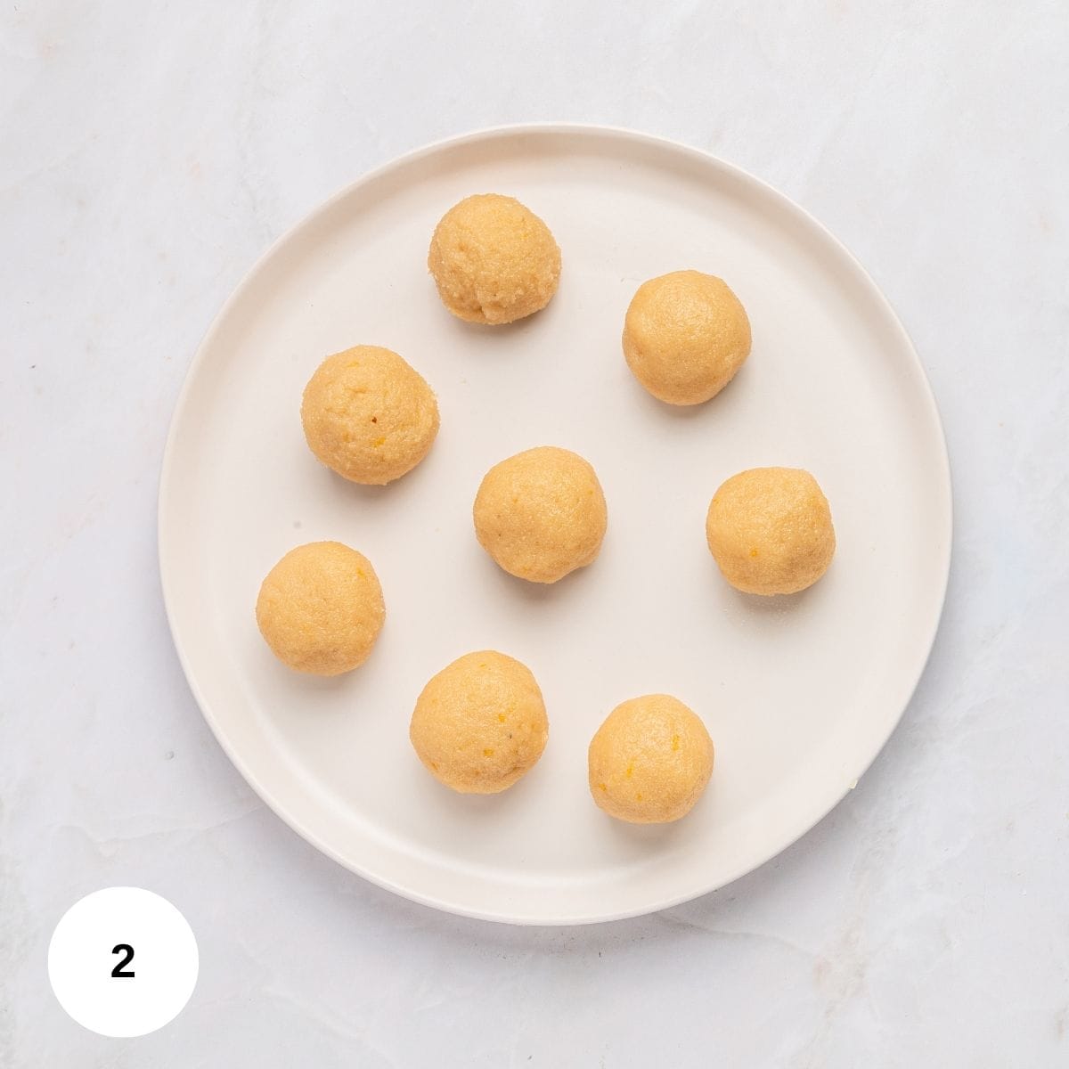 A white plate with eight evenly spaced, round, light golden Italian almond cookies with almond flour sits on a white marble surface. A small white circle with the number 2 is in the bottom left corner.