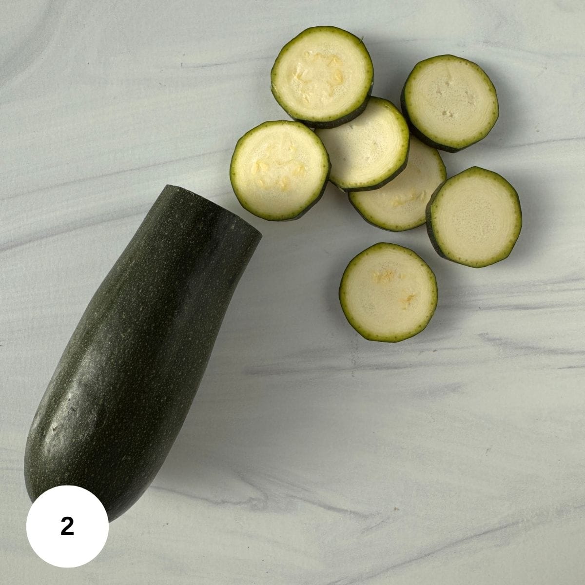 A whole zucchini with several roasted zucchini slices arranged on a light gray marble surface. A white circle with the number 2 is in the bottom left corner.