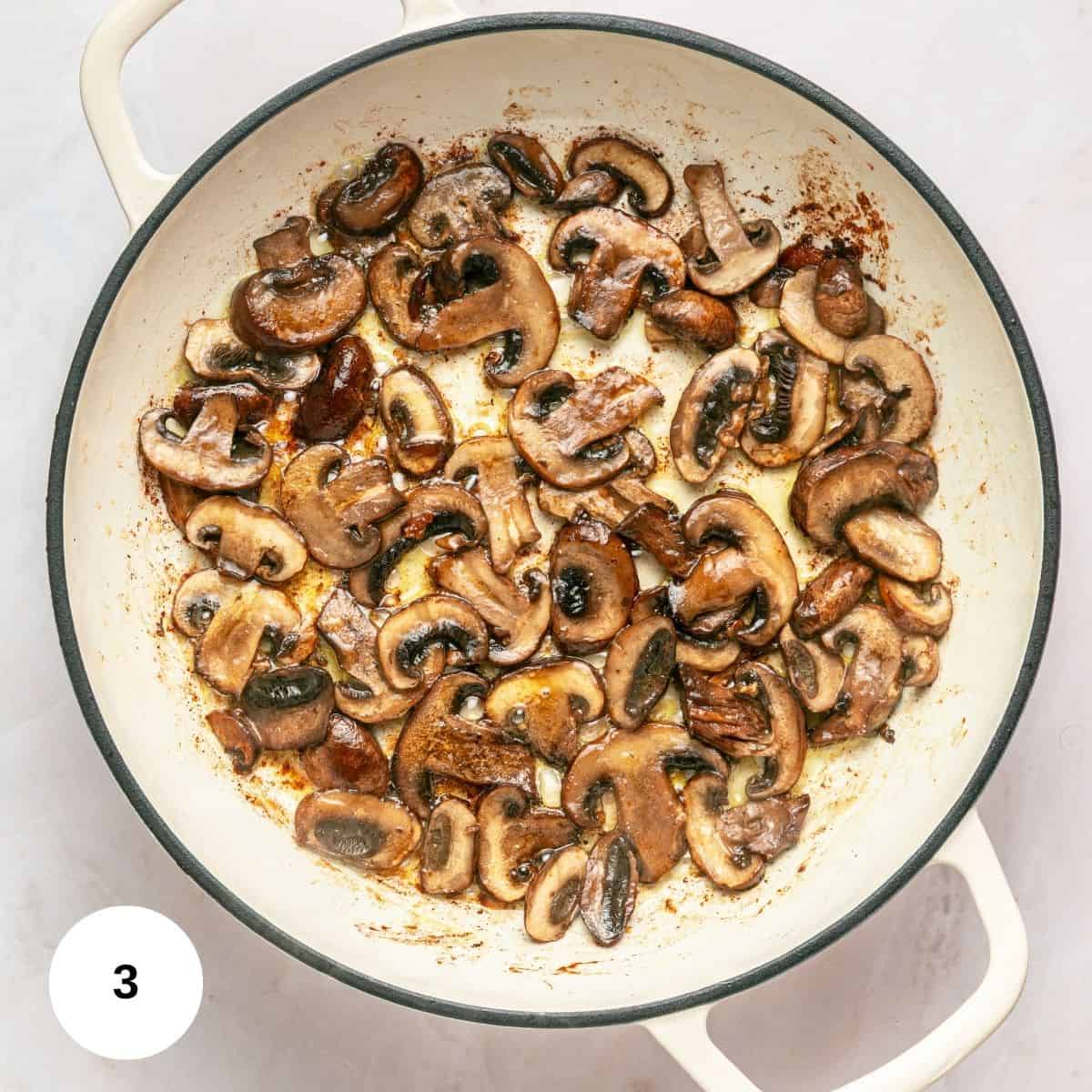 Sliced mushrooms being sautéed in a white enameled pan, showing some browning. This one pan scene highlights the rich flavors developing. The number 3 appears in a white circle at the bottom left corner.