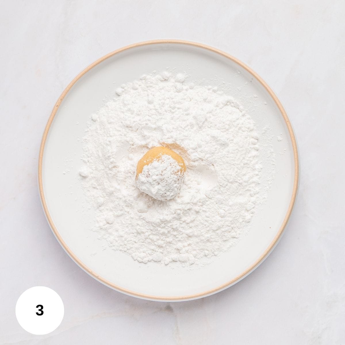 A small ball of dough for Italian almond cookies with almond flour sits on a plate covered with a generous amount of white powdered sugar, ready to be coated. The plate is on a white surface, and a circle with the number 3 is in the lower left corner.