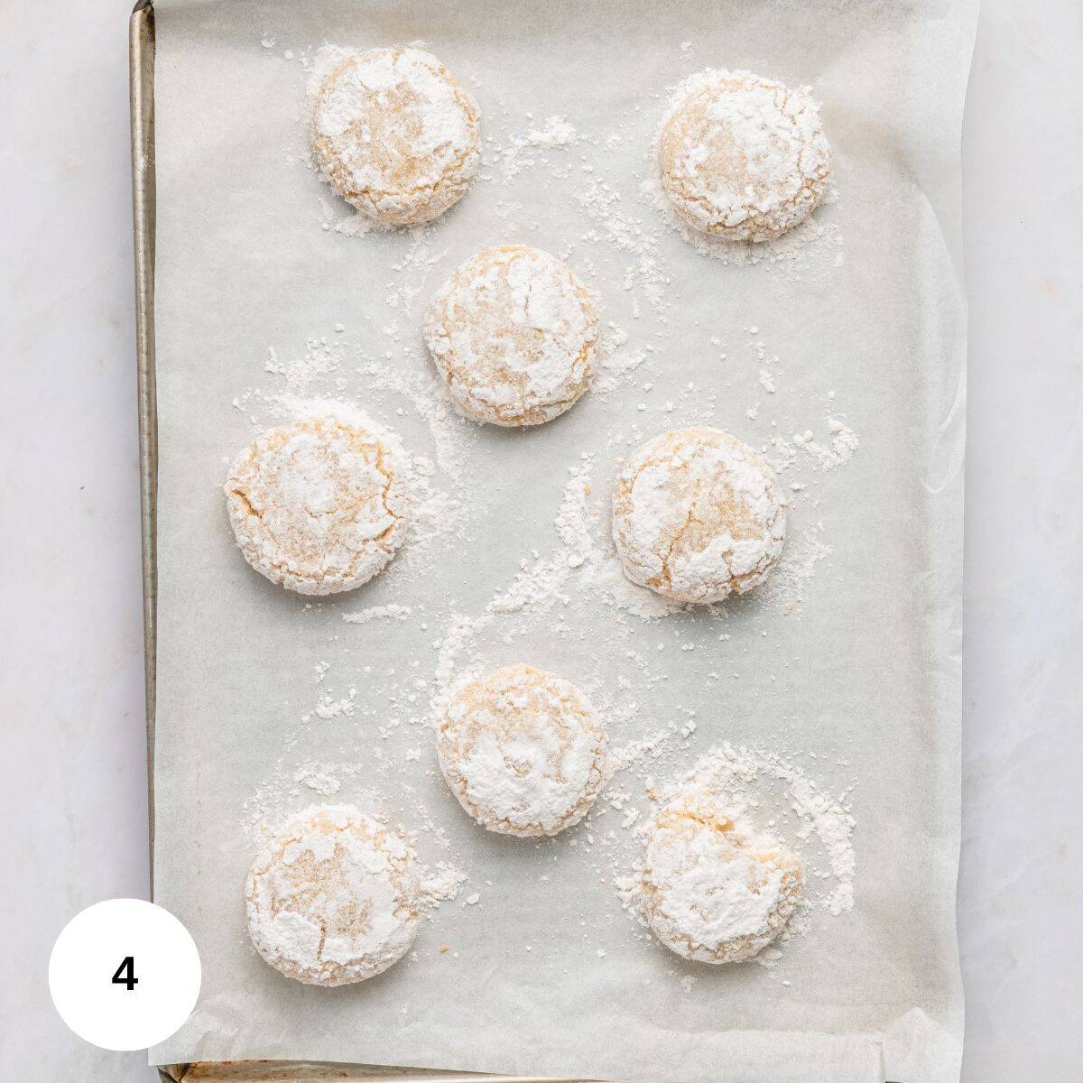 A baking sheet lined with parchment paper holds eight round Italian almond cookies with almond flour, dusted with powdered sugar and spaced evenly apart, ready for baking. A white circle with the number 4 is in the lower left corner.