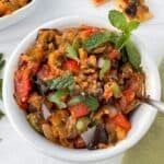 A white bowl filled with colorful Sicilian caponata, featuring chunks of eggplant, tomatoes, bell peppers, and herbs, garnished with fresh mint leaves. A spoon rests in the bowl on a light surface.