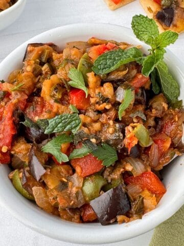 A white bowl filled with colorful Sicilian caponata, featuring chunks of eggplant, tomatoes, bell peppers, and herbs, garnished with fresh mint leaves. A spoon rests in the bowl on a light surface.