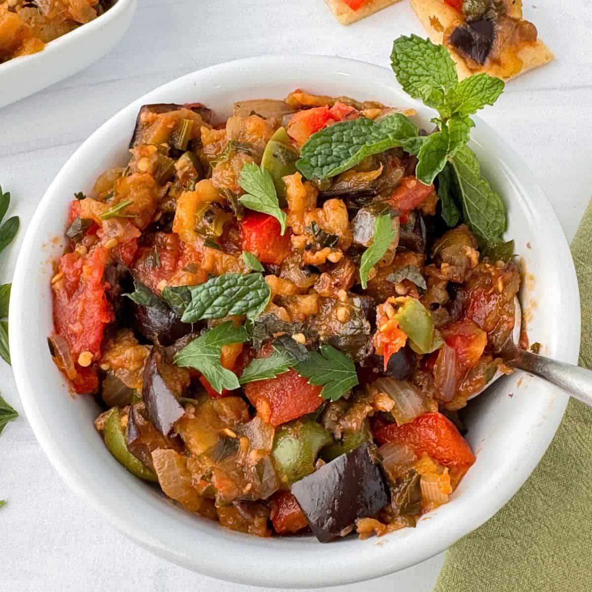 A white bowl filled with colorful Sicilian caponata, featuring chunks of eggplant, tomatoes, bell peppers, and herbs, garnished with fresh mint leaves. A spoon rests in the bowl on a light surface.